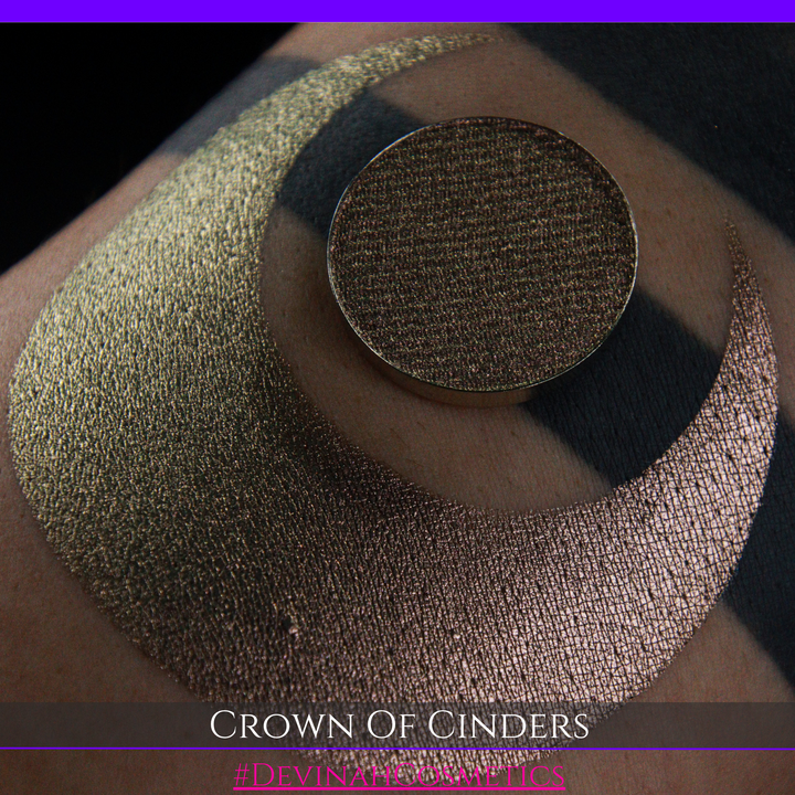 A round eyeshadow pan labeled 'CROWN OF CINDERS' made by Devinah Cosmetics, placed over a large crescent moon swatch of the same shimmering shifty eyeshadow on skin. The eyeshadow has a duochrome multichrome shift between colors bronze, pink, red.