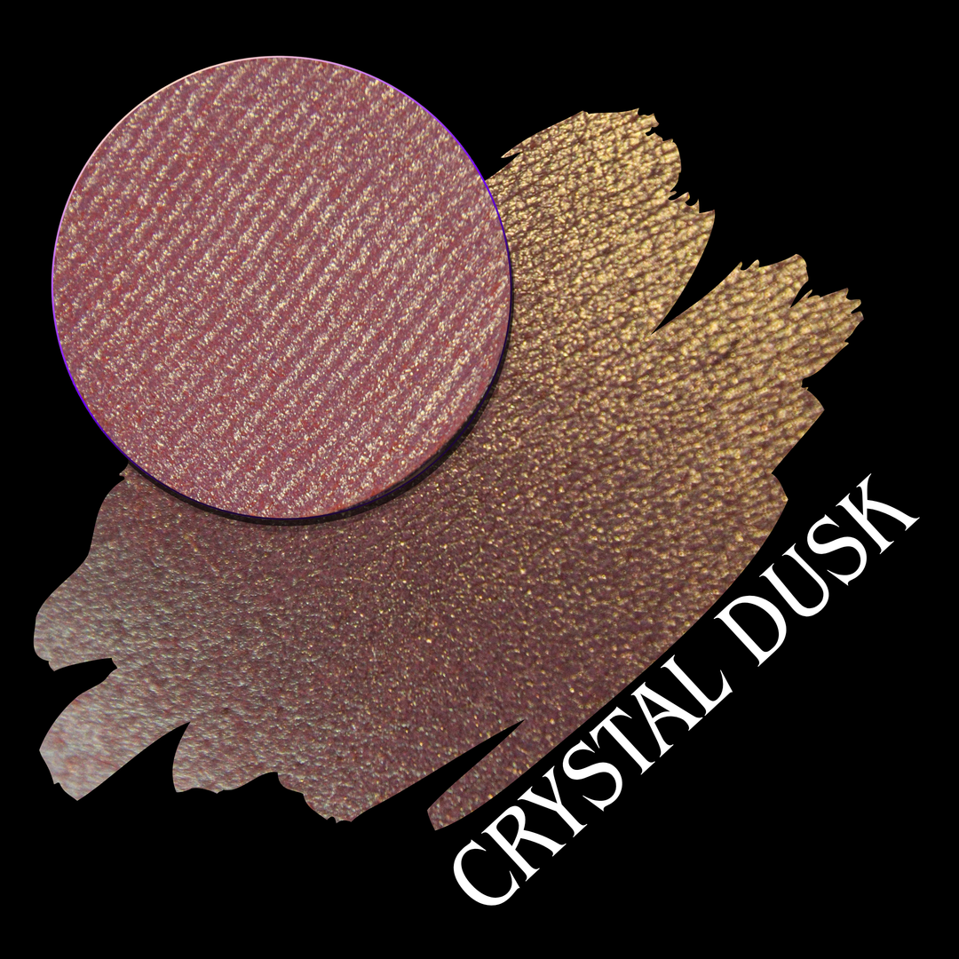 A makeup eyeshadow swatch showing a pink-rose eyeshadow with a golden to green gradient shift with a circular eyeshadow pan on a black background. White text reads 'CRYSTAL DUSK' made by Devinah Cosmetics.