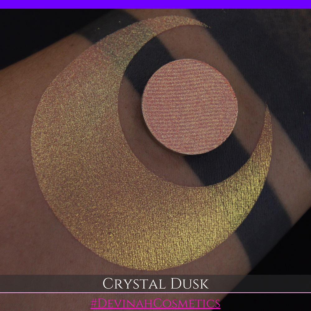 A close-up of a person's arm showing a makeup eyeshadow swatch. The swatch consists of a circular eyeshadow pan placed over a larger crescent-shaped swatch of the same shimmering named Crystal Dusk made by Devinah Cosmetics.