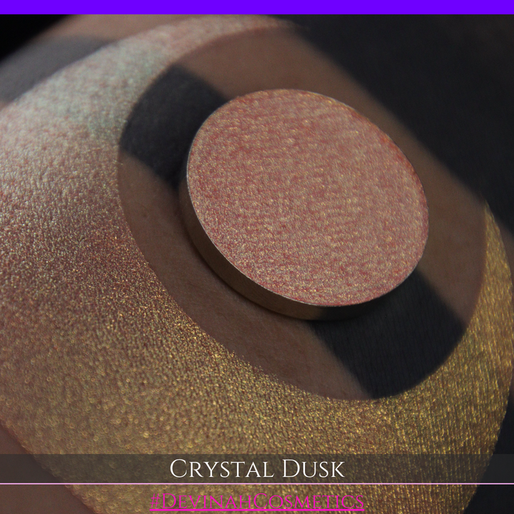 A close-up of a person's arm showing a makeup eyeshadow swatch. The swatch consists of a circular eyeshadow pan placed over a larger crescent-shaped swatch of the same shimmering named Crystal Dusk made by Devinah Cosmetics.