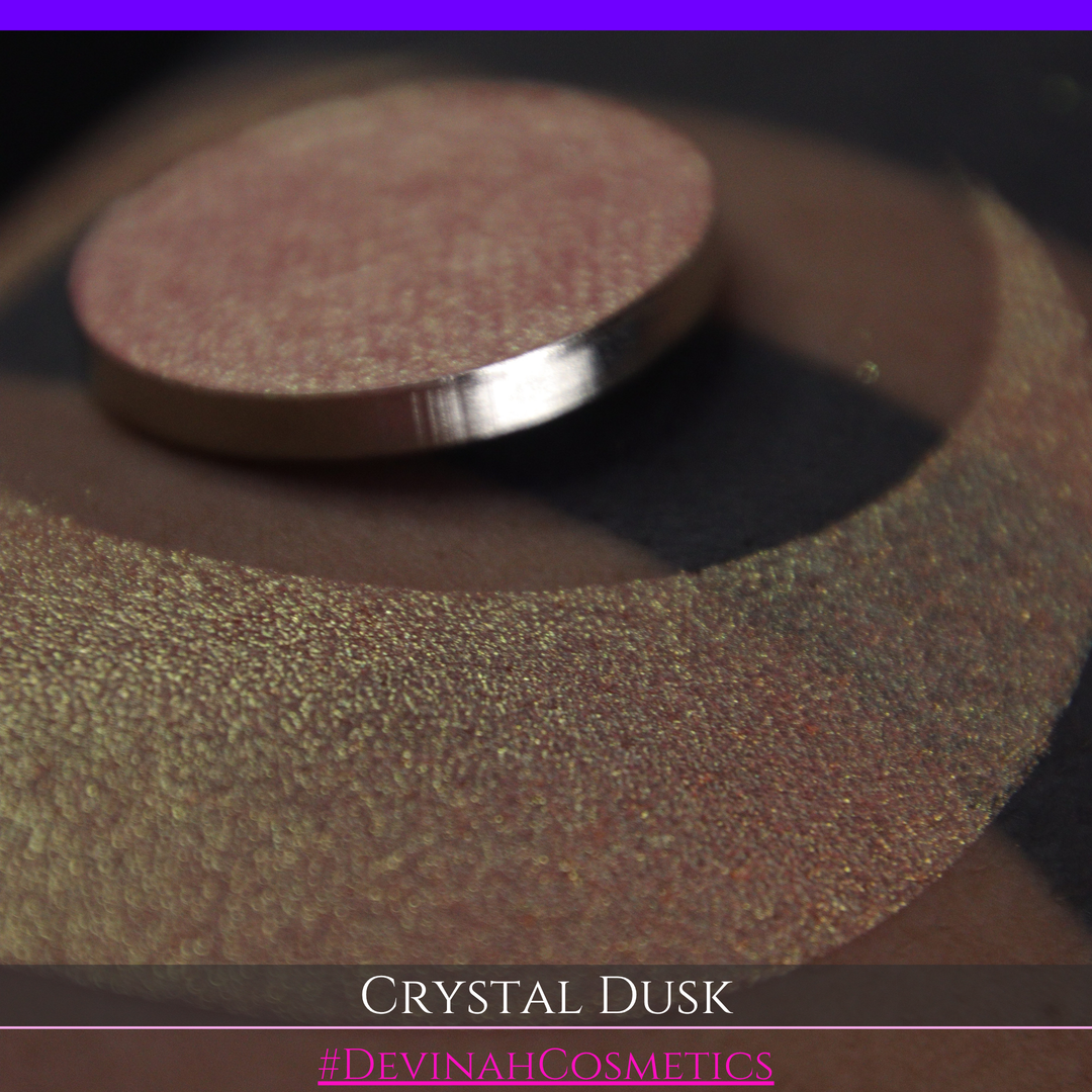 A close-up of a person's arm showing a makeup eyeshadow swatch. The swatch consists of a circular eyeshadow pan placed over a larger crescent-shaped swatch of the same shimmering named Crystal Dusk made by Devinah Cosmetics.