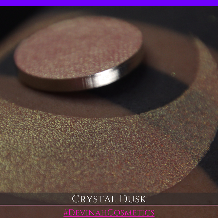 A close-up of a person's arm showing a makeup eyeshadow swatch. The swatch consists of a circular eyeshadow pan placed over a larger crescent-shaped swatch of the same shimmering named Crystal Dusk made by Devinah Cosmetics.