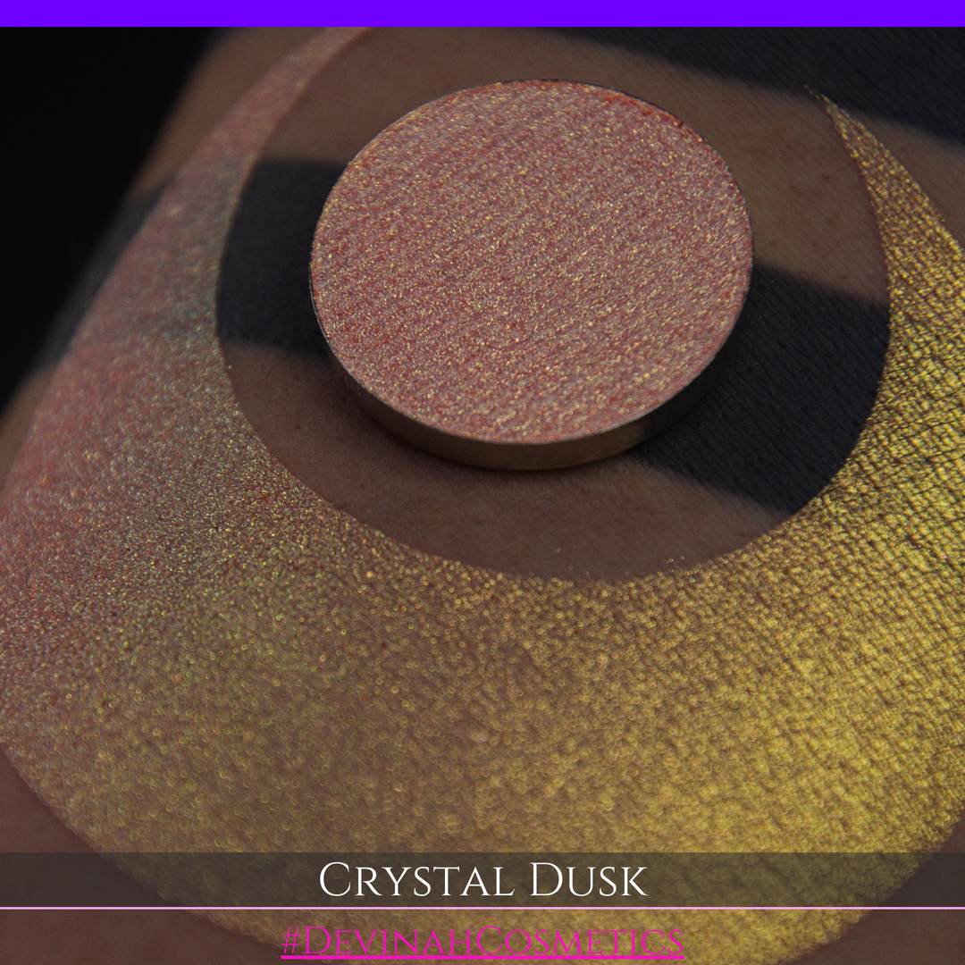 A close-up of a person's arm showing a makeup eyeshadow swatch. The swatch consists of a circular eyeshadow pan placed over a larger crescent-shaped swatch of the same shimmering named Crystal Dusk made by Devinah Cosmetics.