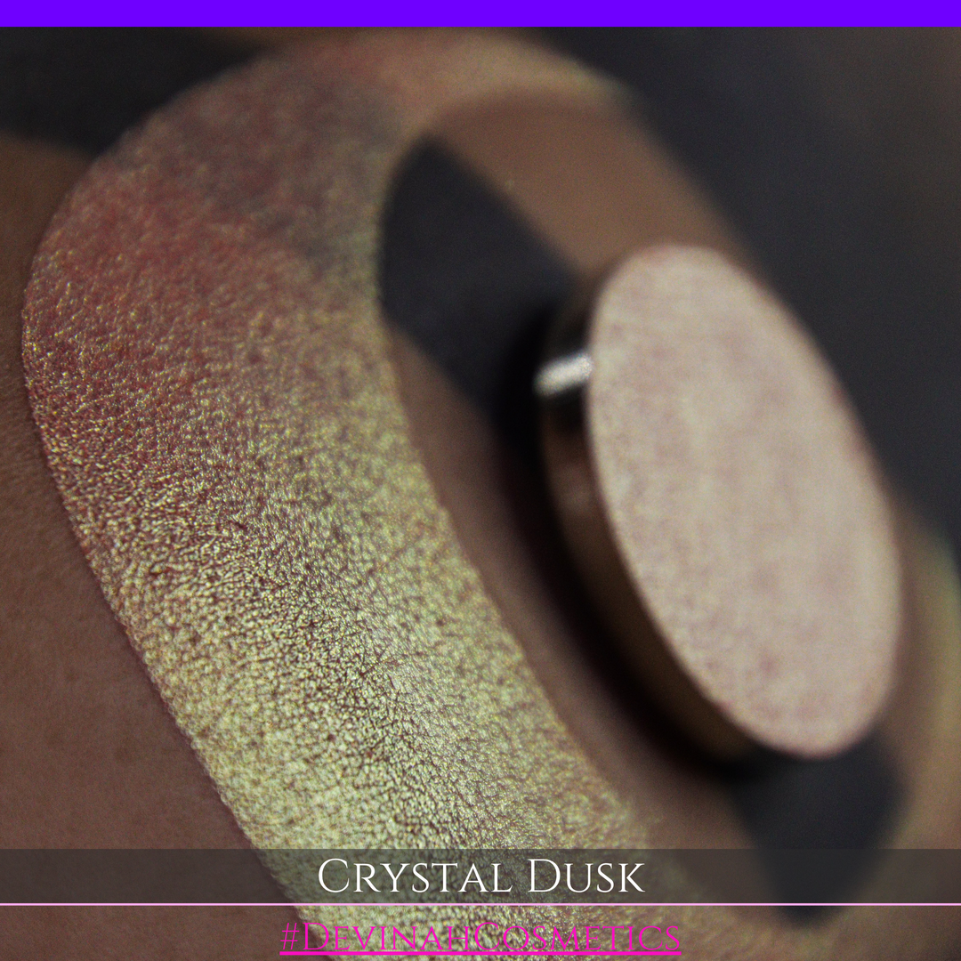 A close-up of a person's arm showing a makeup eyeshadow swatch. The swatch consists of a circular eyeshadow pan placed over a larger crescent-shaped swatch of the same shimmering named Crystal Dusk made by Devinah Cosmetics.