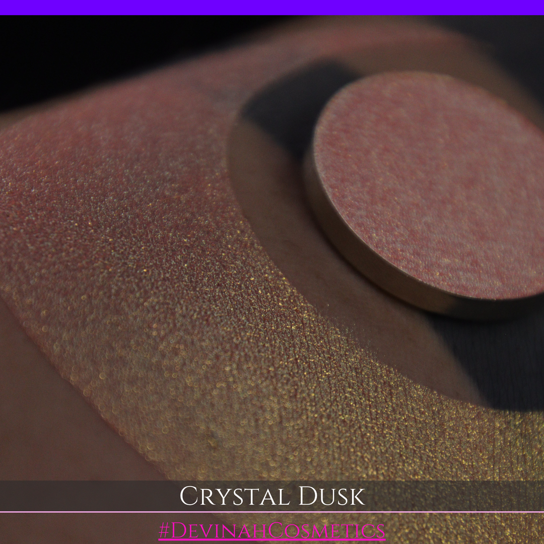 A close-up of a person's arm showing a makeup eyeshadow swatch. The swatch consists of a circular eyeshadow pan placed over a larger crescent-shaped swatch of the same shimmering named Crystal Dusk made by Devinah Cosmetics.