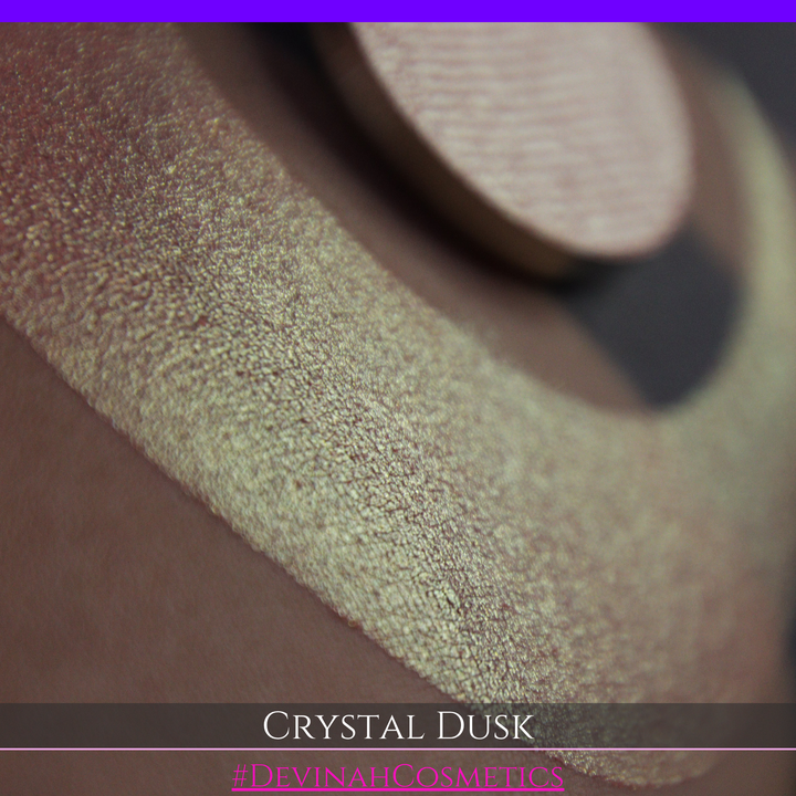 A close-up of a person's arm showing a makeup eyeshadow swatch. The swatch consists of a circular eyeshadow pan placed over a larger crescent-shaped swatch of the same shimmering named Crystal Dusk made by Devinah Cosmetics.