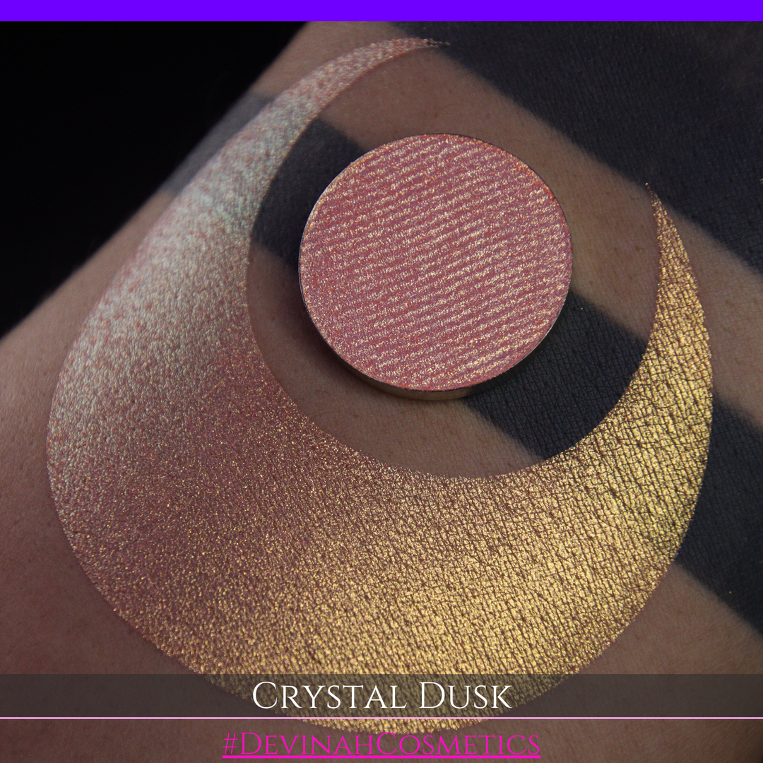 A close-up of a person's arm showing a makeup eyeshadow swatch. The swatch consists of a circular eyeshadow pan placed over a larger crescent-shaped swatch of the same shimmering named Crystal Dusk made by Devinah Cosmetics.