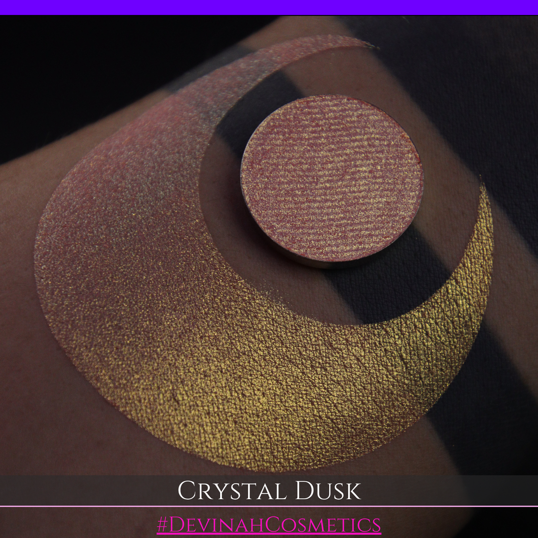 A close-up of a person's arm showing a makeup eyeshadow swatch. The swatch consists of a circular eyeshadow pan placed over a larger crescent-shaped swatch of the same shimmering named Crystal Dusk made by Devinah Cosmetics.