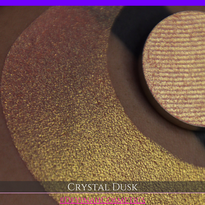 A close-up of a person's arm showing a makeup eyeshadow swatch. The swatch consists of a circular eyeshadow pan placed over a larger crescent-shaped swatch of the same shimmering named Crystal Dusk made by Devinah Cosmetics.