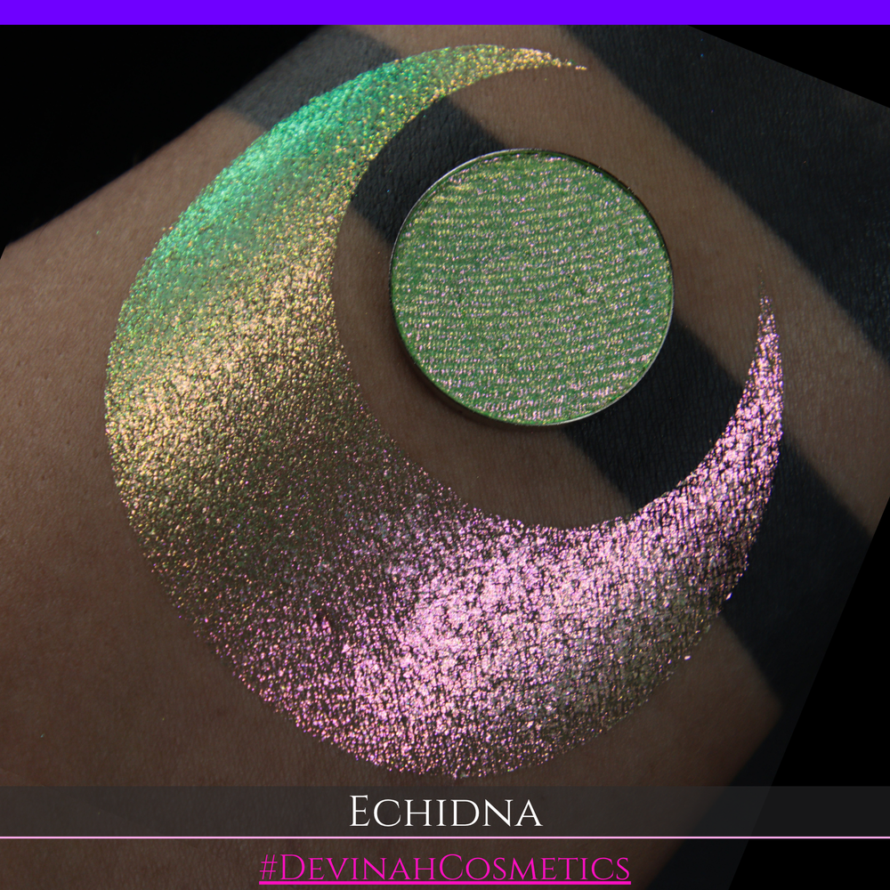 A round eyeshadow pan labeled 'ECHIDNA' made by Devinah Cosmetics, placed over a large crescent moon swatch of the same shimmering green-to-pink multichrome eyeshadow on skin. The eyeshadow has an iridescent, metallic finish shifting between  colors green, gold, and pink.