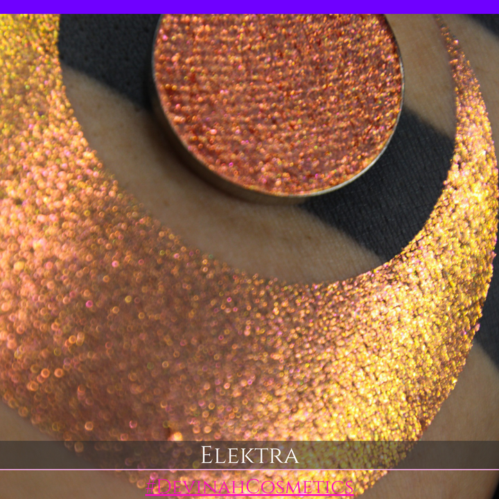 A round eyeshadow pan labeled 'ELEKTRA' made by Devinah Cosmetics, placed over a large crescent moon swatch of the same shimmering shifty duochrome multichrome eyeshadow on skin. The eyeshadow has a shift between colors green, gold, pink.