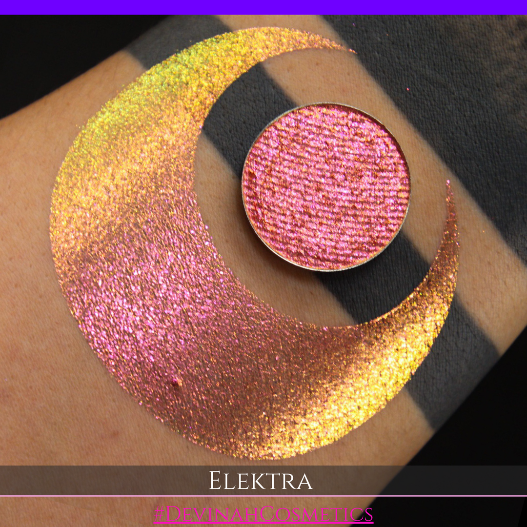 A round eyeshadow pan labeled 'ELEKTRA' made by Devinah Cosmetics, placed over a large crescent moon swatch of the same shimmering shifty duochrome multichrome eyeshadow on skin. The eyeshadow has a shift between colors green, gold, pink.