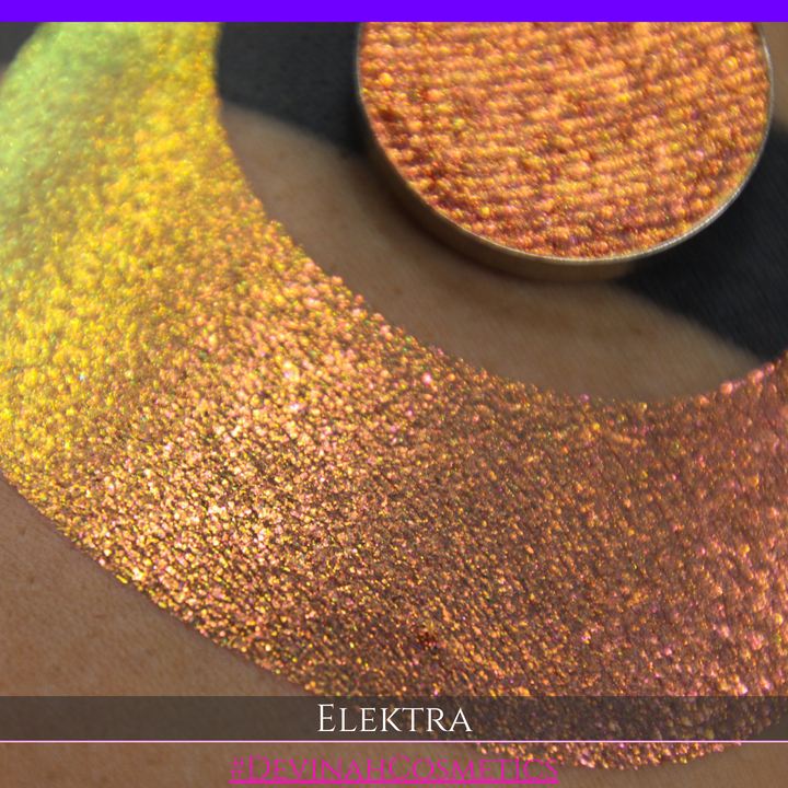 A round eyeshadow pan labeled 'ELEKTRA' made by Devinah Cosmetics, placed over a large crescent moon swatch of the same shimmering shifty duochrome multichrome eyeshadow on skin. The eyeshadow has a shift between colors green, gold, pink.