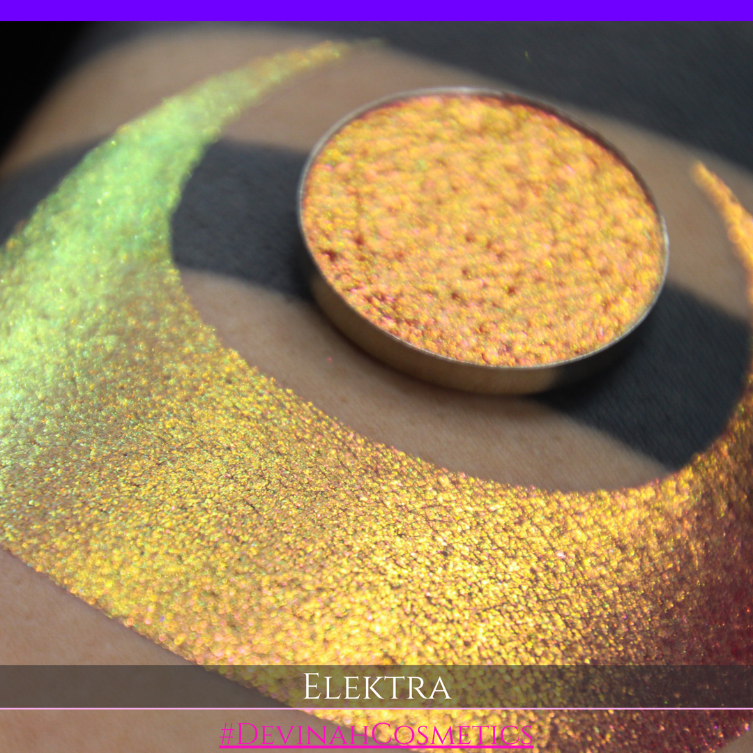 A round eyeshadow pan labeled 'ELEKTRA' made by Devinah Cosmetics, placed over a large crescent moon swatch of the same shimmering shifty duochrome multichrome eyeshadow on skin. The eyeshadow has a shift between colors green, gold, pink.