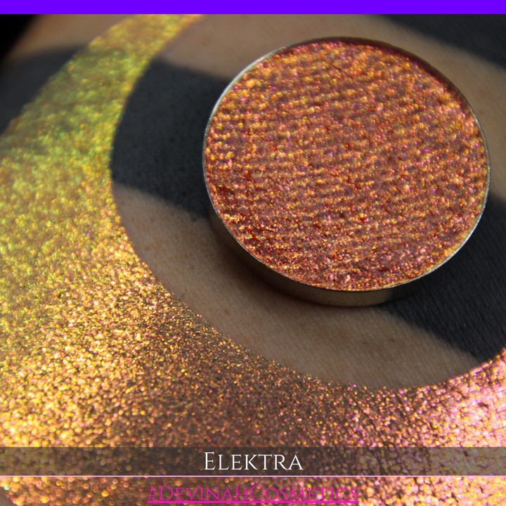 A round eyeshadow pan labeled 'ELEKTRA' made by Devinah Cosmetics, placed over a large crescent moon swatch of the same shimmering shifty duochrome multichrome eyeshadow on skin. The eyeshadow has a shift between colors green, gold, pink.