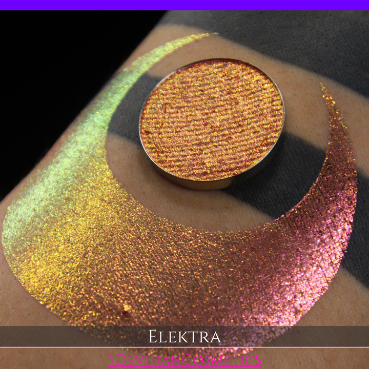 A round eyeshadow pan labeled 'ELEKTRA' made by Devinah Cosmetics, placed over a large crescent moon swatch of the same shimmering shifty duochrome multichrome eyeshadow on skin. The eyeshadow has a shift between colors green, gold, pink.