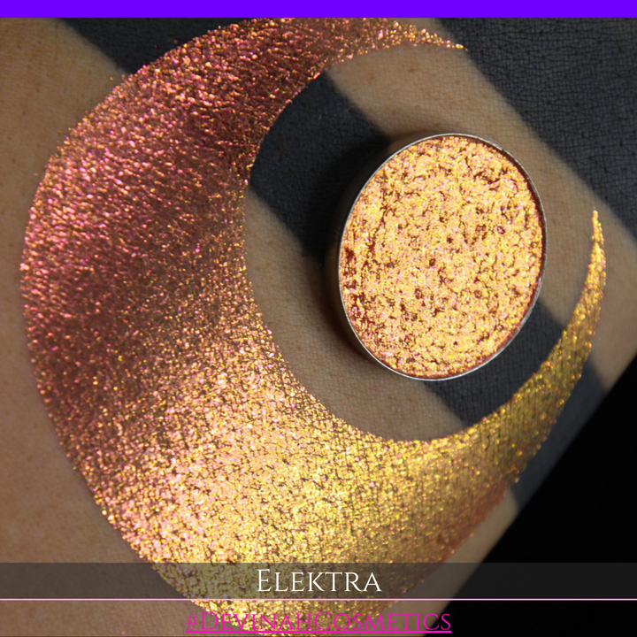 A round eyeshadow pan labeled 'ELEKTRA' made by Devinah Cosmetics, placed over a large crescent moon swatch of the same shimmering shifty duochrome multichrome eyeshadow on skin. The eyeshadow has a shift between colors green, gold, pink.