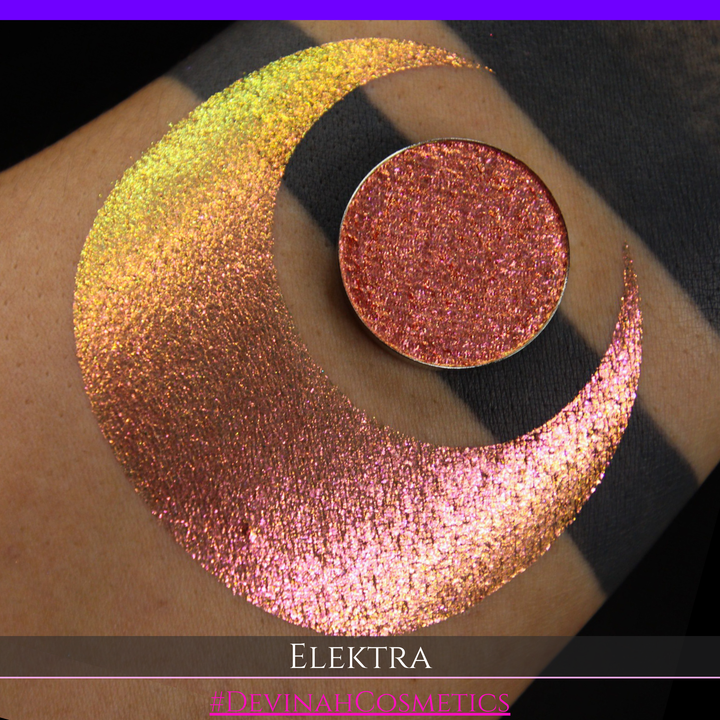 A round eyeshadow pan labeled 'ELEKTRA' made by Devinah Cosmetics, placed over a large crescent moon swatch of the same shimmering shifty duochrome multichrome eyeshadow on skin. The eyeshadow has a shift between colors green, gold, pink.
