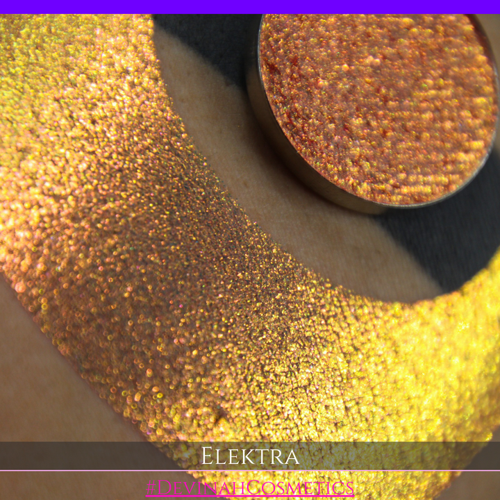 A round eyeshadow pan labeled 'ELEKTRA' made by Devinah Cosmetics, placed over a large crescent moon swatch of the same shimmering shifty duochrome multichrome eyeshadow on skin. The eyeshadow has a shift between colors green, gold, pink.