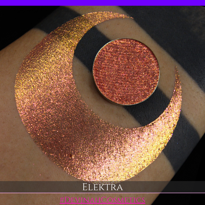 A round eyeshadow pan labeled 'ELEKTRA' made by Devinah Cosmetics, placed over a large crescent moon swatch of the same shimmering shifty duochrome multichrome eyeshadow on skin. The eyeshadow has a shift between colors green, gold, pink.