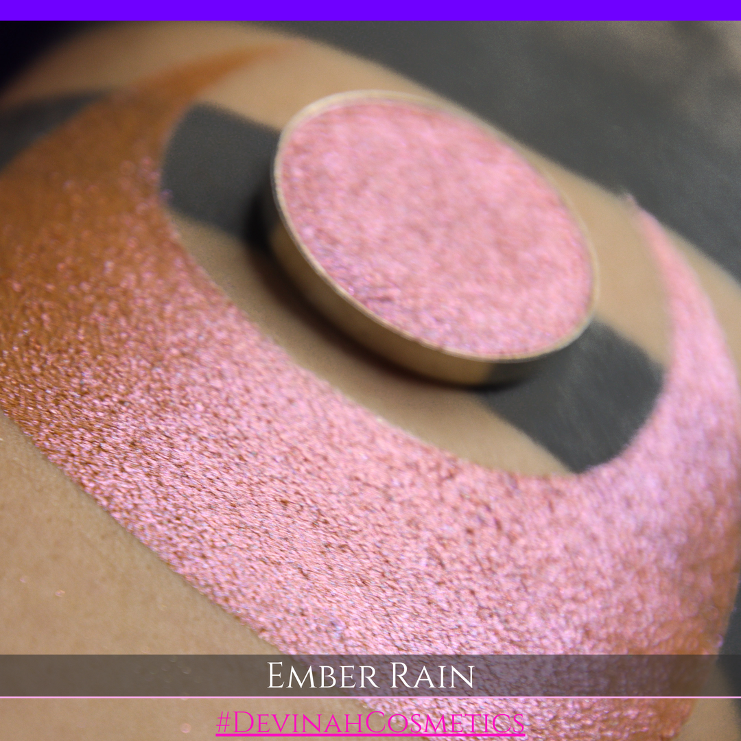 A close-up of a person's skin showing a large swatch of shimmering eyeshadow in a crescent shape, with a round makeup pan placed on top. Text at the bottom reads 'EMBER RAIN' and '#DEVINAH COSMETICS'.