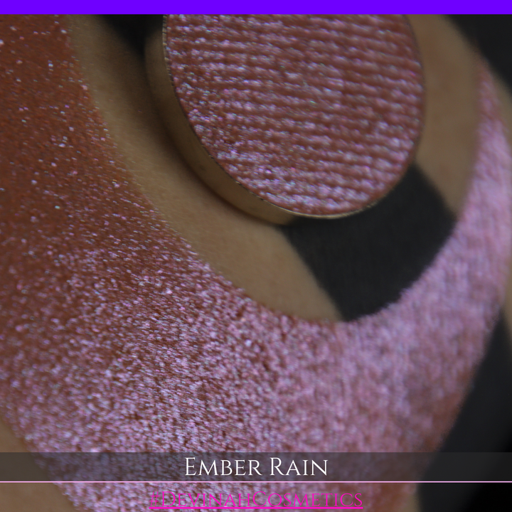 A close-up of a person's skin showing a large swatch of shimmering eyeshadow in a crescent shape, with a round makeup pan placed on top. Text at the bottom reads 'EMBER RAIN' and '#DEVINAH COSMETICS'.