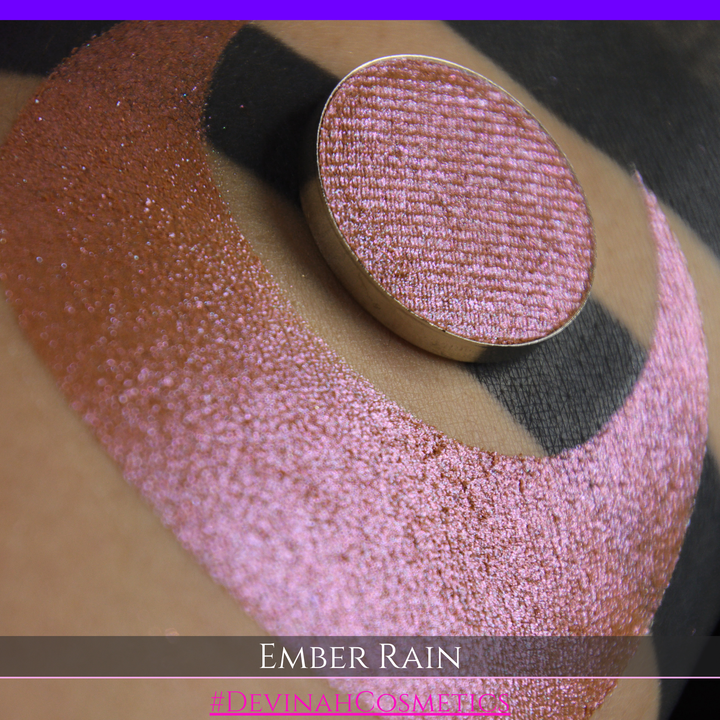 A close-up of a person's skin showing a large swatch of shimmering eyeshadow in a crescent shape, with a round makeup pan placed on top. Text at the bottom reads 'EMBER RAIN' and '#DEVINAH COSMETICS'.