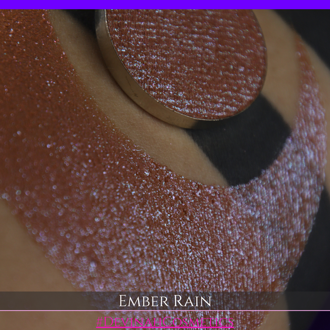 A close-up of a person's skin showing a large swatch of shimmering eyeshadow in a crescent shape, with a round makeup pan placed on top. Text at the bottom reads 'EMBER RAIN' and '#DEVINAH COSMETICS'.