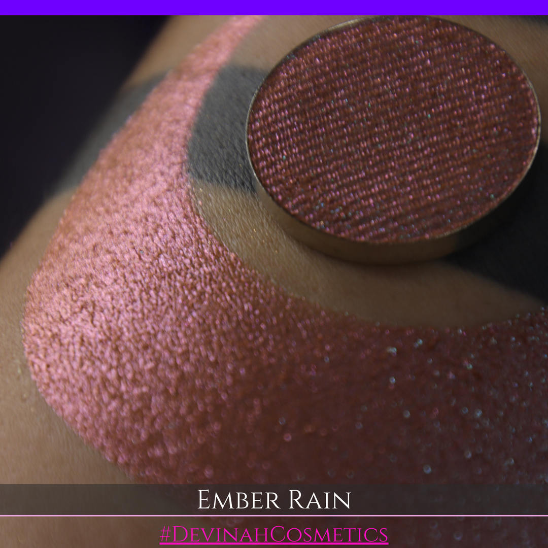A close-up of a person's skin showing a large swatch of shimmering eyeshadow in a crescent shape, with a round makeup pan placed on top. Text at the bottom reads 'EMBER RAIN' and '#DEVINAH COSMETICS'.