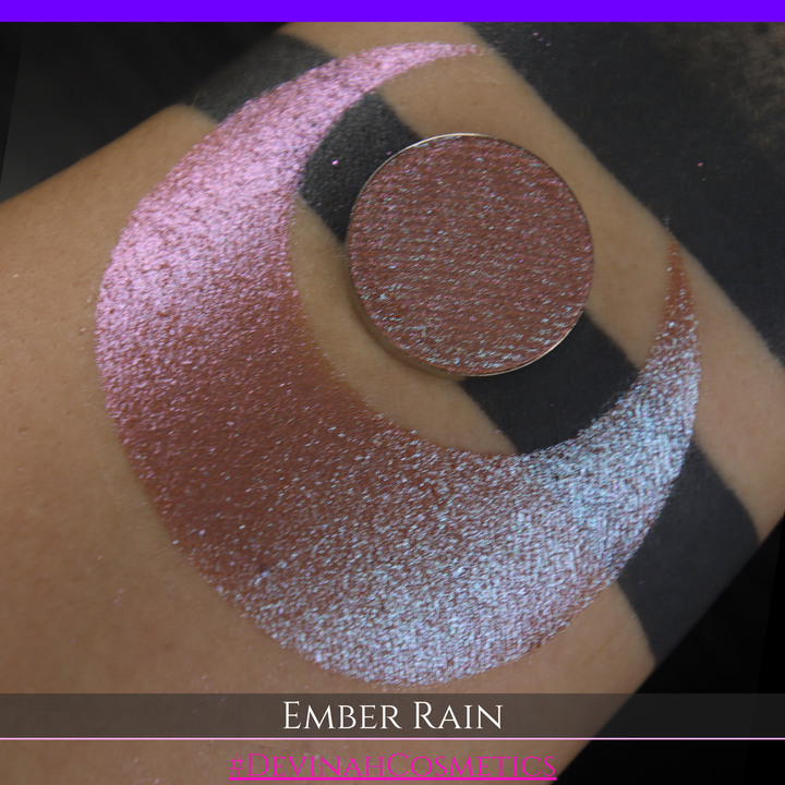 A close-up of a person's skin showing a large swatch of shimmering eyeshadow in a crescent shape, with a round makeup pan placed on top. Text at the bottom reads 'EMBER RAIN' and '#DEVINAH COSMETICS'.