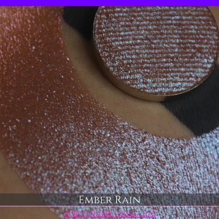A close-up of a person's skin showing a large swatch of shimmering eyeshadow in a crescent shape, with a round makeup pan placed on top. Text at the bottom reads 'EMBER RAIN' and '#DEVINAH COSMETICS'.