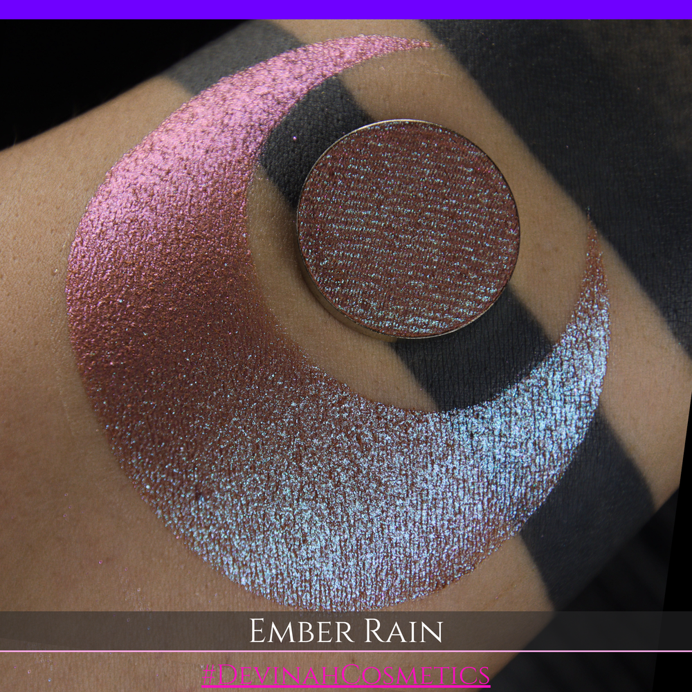 A close-up of a person's skin showing a large swatch of shimmering eyeshadow in a crescent shape, with a round makeup pan placed on top. Text at the bottom reads 'EMBER RAIN' and '#DEVINAH COSMETICS'.