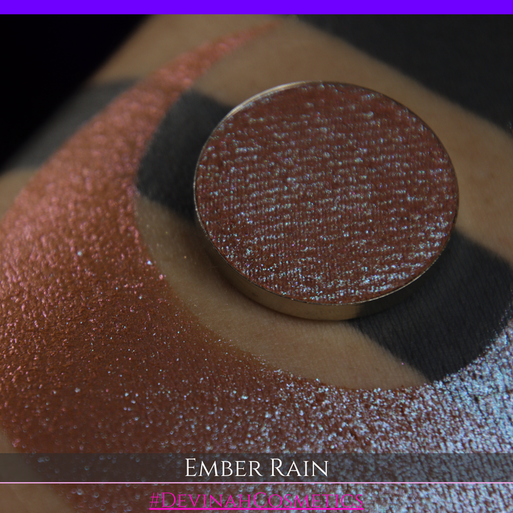 A close-up of a person's skin showing a large swatch of shimmering eyeshadow in a crescent shape, with a round makeup pan placed on top. Text at the bottom reads 'EMBER RAIN' and '#DEVINAH COSMETICS'.