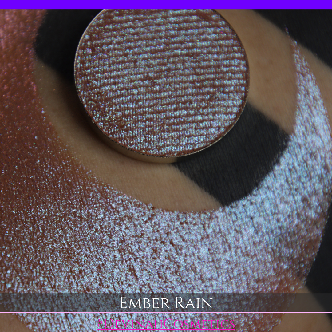 A close-up of a person's skin showing a large swatch of shimmering eyeshadow in a crescent shape, with a round makeup pan placed on top. Text at the bottom reads 'EMBER RAIN' and '#DEVINAH COSMETICS'.