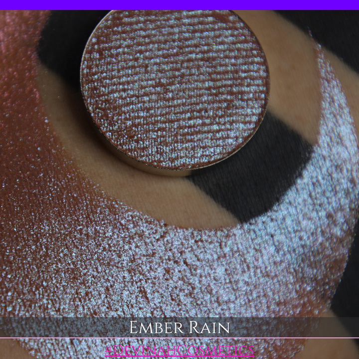 A close-up of a person's skin showing a large swatch of shimmering eyeshadow in a crescent shape, with a round makeup pan placed on top. Text at the bottom reads 'EMBER RAIN' and '#DEVINAH COSMETICS'.