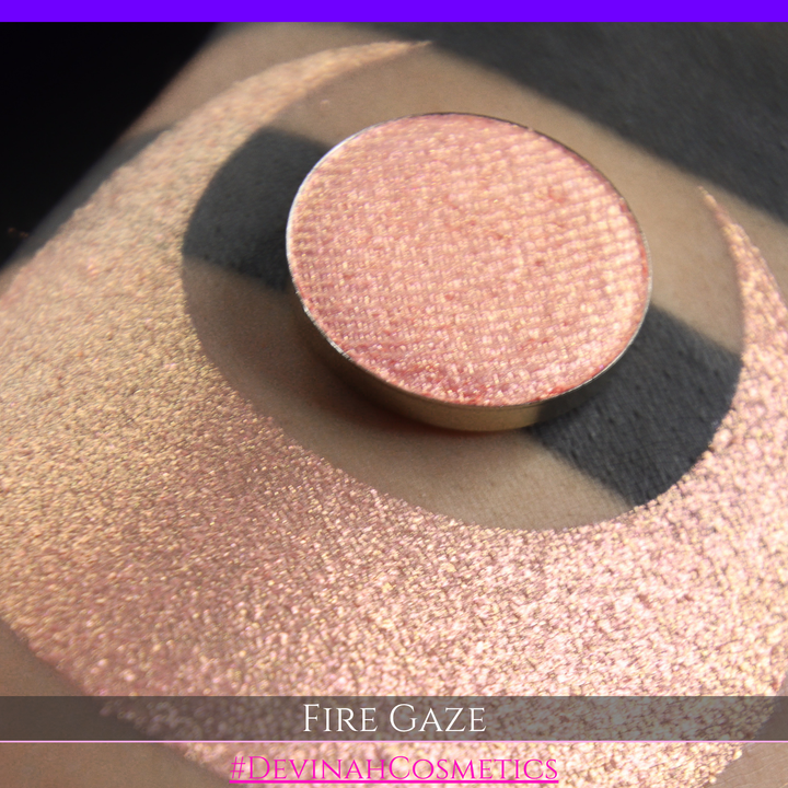 A makeup color swatch showing a shimmering pink-rose eyeshadow with a circular eyeshadow pan and the text 'FIRE GAZE' on a black background made by Devinah Cosmetics.