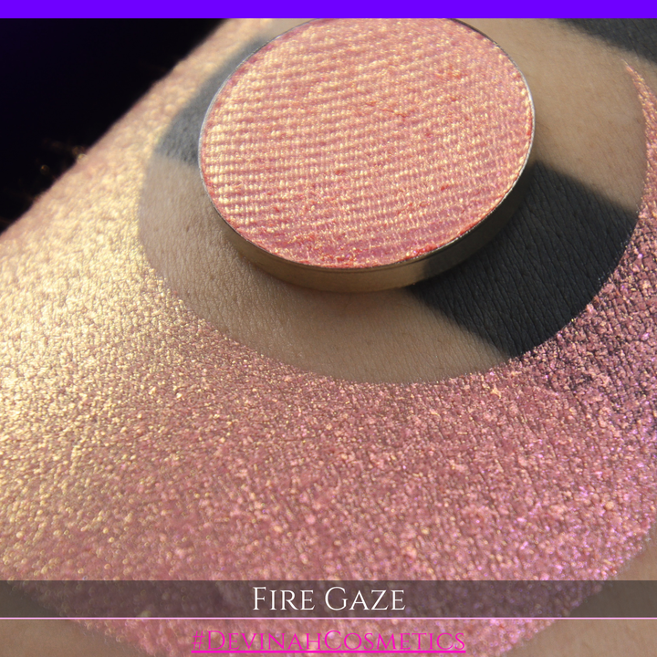 A makeup color swatch showing a shimmering pink-rose eyeshadow with a circular eyeshadow pan and the text 'FIRE GAZE' on a black background made by Devinah Cosmetics.