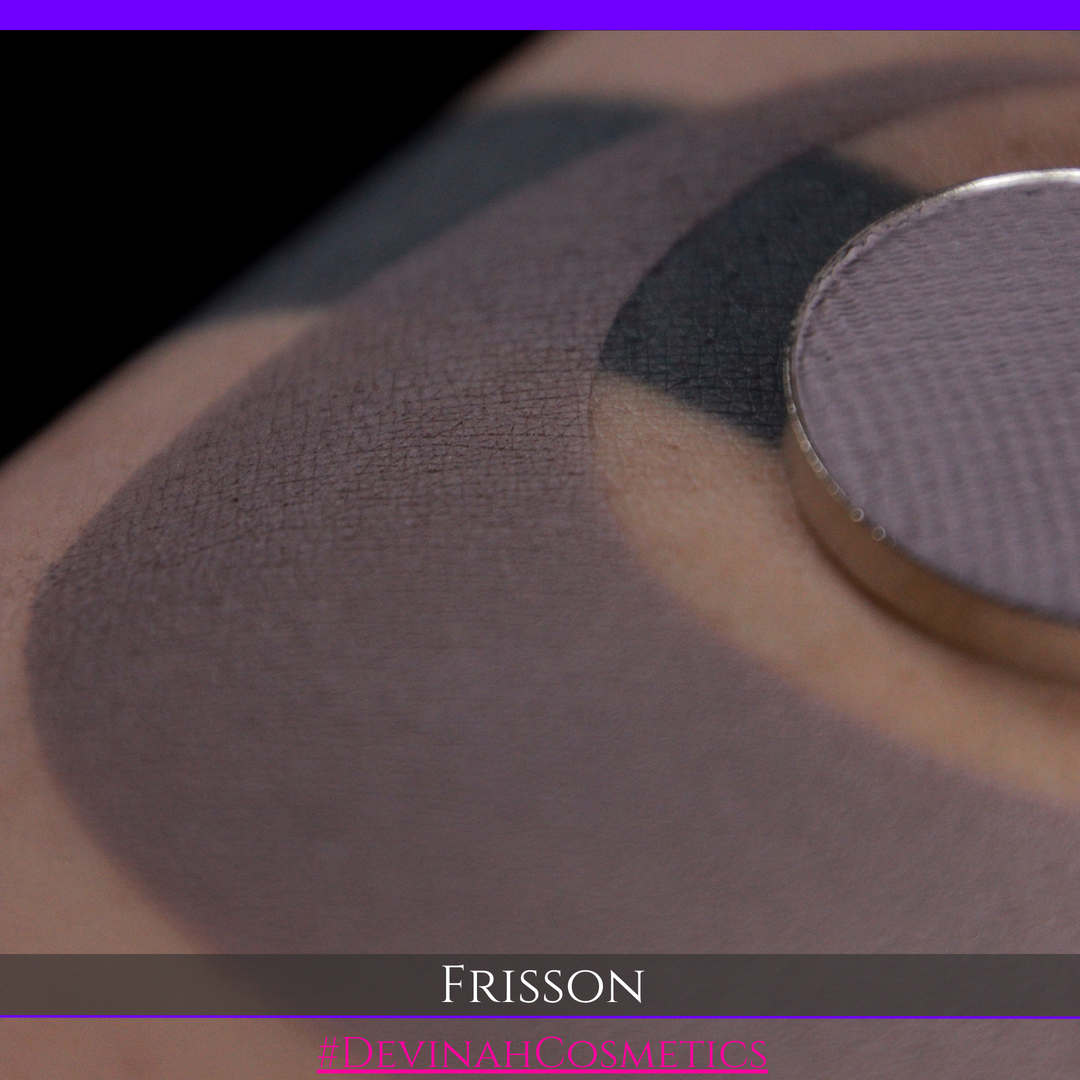 Close-up of a makeup swatch showing a mauve-gray eyeshadow applied on skin, with a round pressed powder pan in the upper right and the text 'FRISSON' and 'DEVINAH COSMETICS' at the bottom.