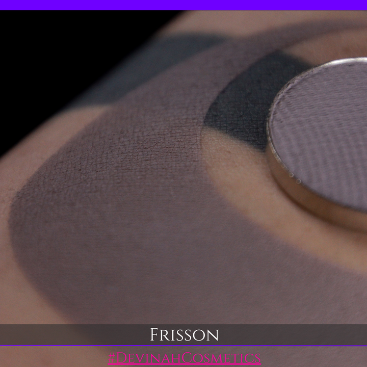 Close-up of a makeup swatch showing a mauve-gray eyeshadow applied on skin, with a round pressed powder pan in the upper right and the text 'FRISSON' and 'DEVINAH COSMETICS' at the bottom.