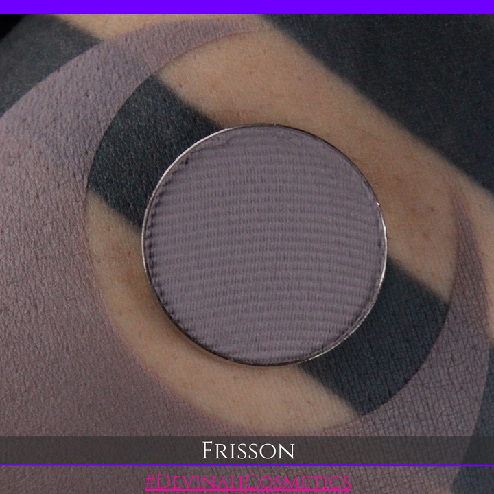A close-up of a round eyeshadow pan filled with a muted mauve-gray powder, placed over circular swatches of the same shade on skin. Text at the bottom reads 'FRISSON' and '#DEVINAH COSMETICS'.