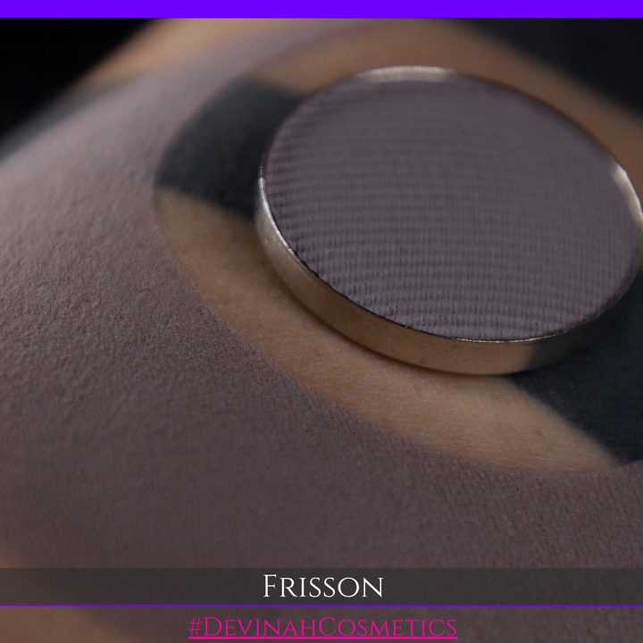 A close-up of a round makeup pan containing a dark gray-purple pressed powder, resting on skin. The bottom of the image features a dark banner with the brand name 'FRISSON' and the text '#DEVINAHCOSMETICS'.
