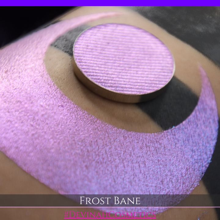 A close-up of skin with a large iridescent pink-blue crescent-shaped makeup swatch and a matching circulareyeshadow pan, with text reading 'FROST BANE' and '#DEVINAH COSMETICS' at the bottom.