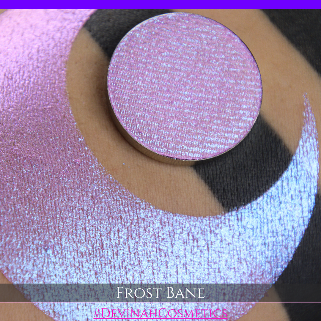 A close-up of skin with a large iridescent pink-blue crescent-shaped makeup swatch and a matching circulareyeshadow pan, with text reading 'FROST BANE' and '#DEVINAH COSMETICS' at the bottom.
