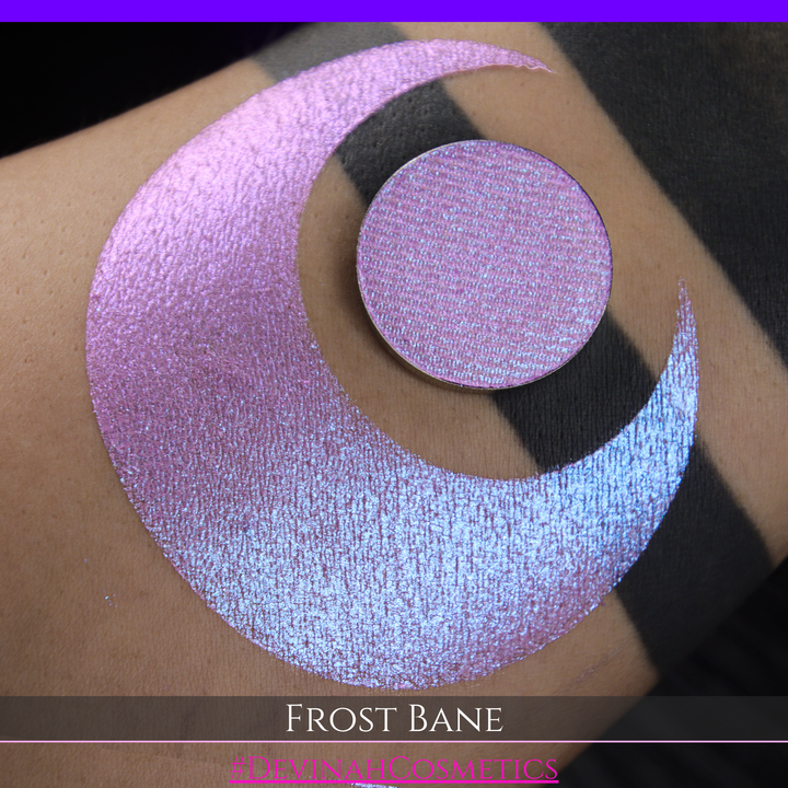 A close-up of skin with a large iridescent pink-blue crescent-shaped makeup swatch and a matching circulareyeshadow pan, with text reading 'FROST BANE' and '#DEVINAH COSMETICS' at the bottom.