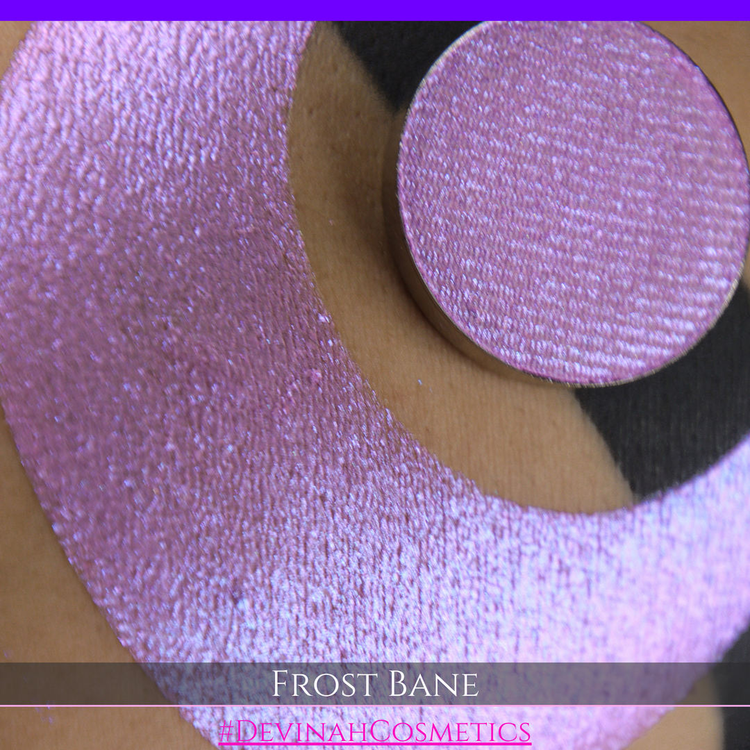 A close-up of skin with a large iridescent pink-blue crescent-shaped makeup swatch and a matching circulareyeshadow pan, with text reading 'FROST BANE' and '#DEVINAH COSMETICS' at the bottom.