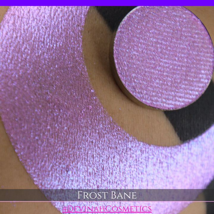 A close-up of skin with a large iridescent pink-blue crescent-shaped makeup swatch and a matching circulareyeshadow pan, with text reading 'FROST BANE' and '#DEVINAH COSMETICS' at the bottom.