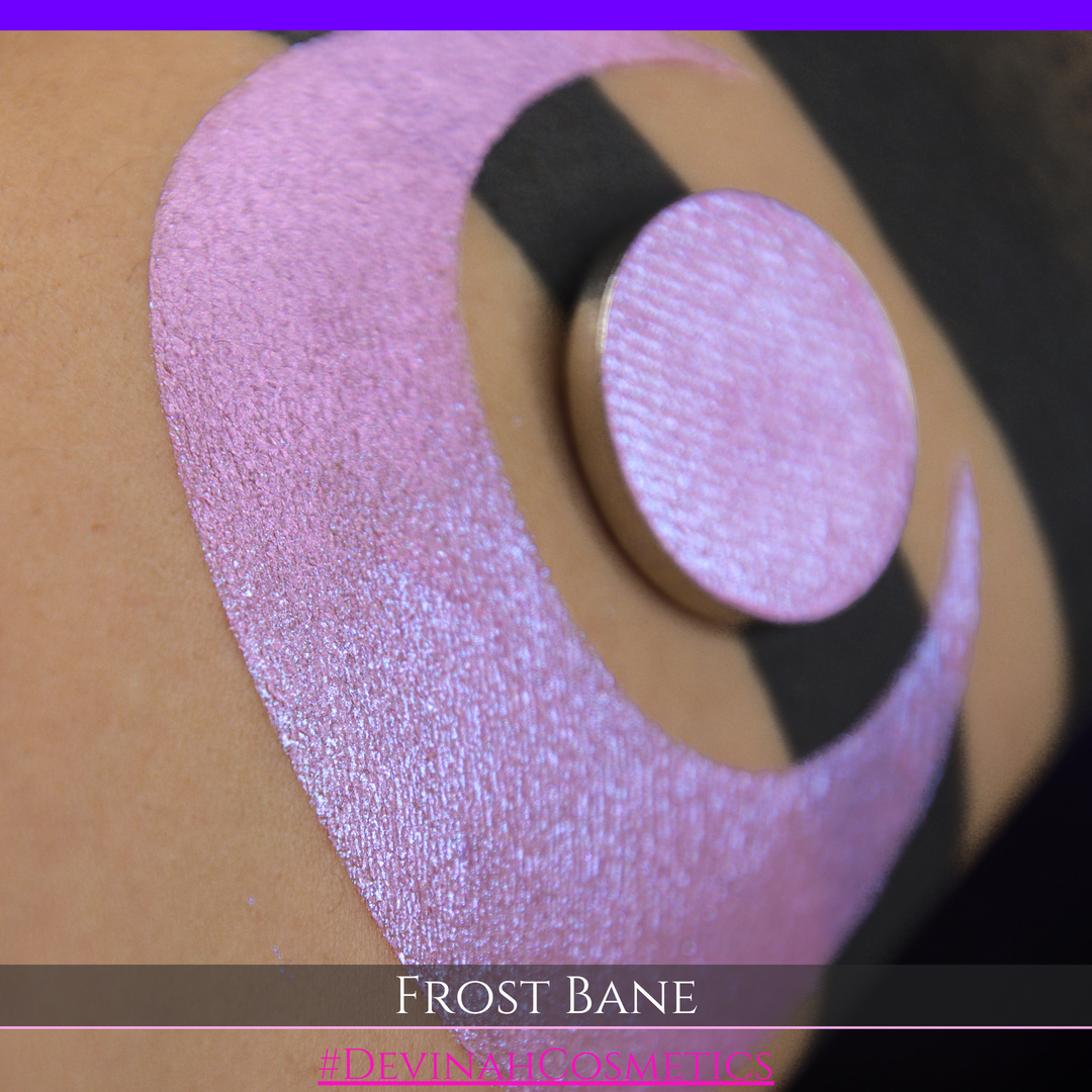 A close-up of skin with a large iridescent pink-blue crescent-shaped makeup swatch and a matching circulareyeshadow pan, with text reading 'FROST BANE' and '#DEVINAH COSMETICS' at the bottom.