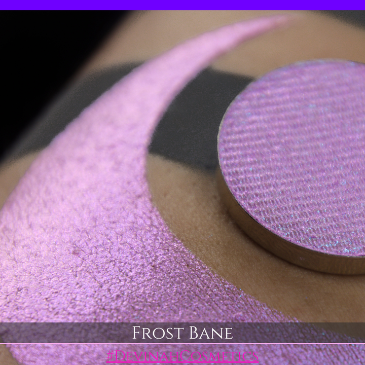 A close-up of skin with a large iridescent pink-blue crescent-shaped makeup swatch and a matching circulareyeshadow pan, with text reading 'FROST BANE' and '#DEVINAH COSMETICS' at the bottom.