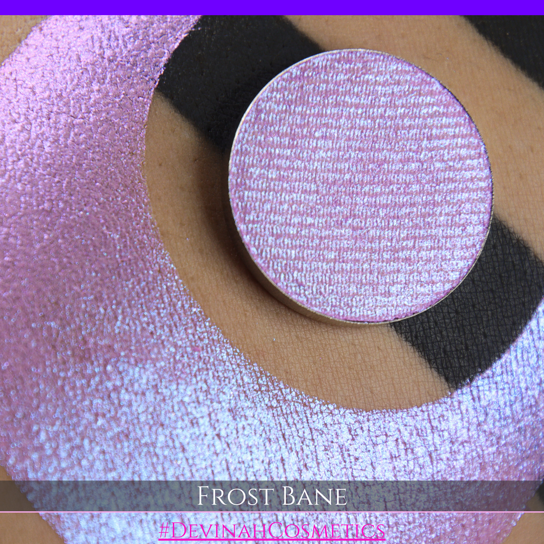 A close-up of skin with a large iridescent pink-blue crescent-shaped makeup swatch and a matching circulareyeshadow pan, with text reading 'FROST BANE' and '#DEVINAH COSMETICS' at the bottom.
