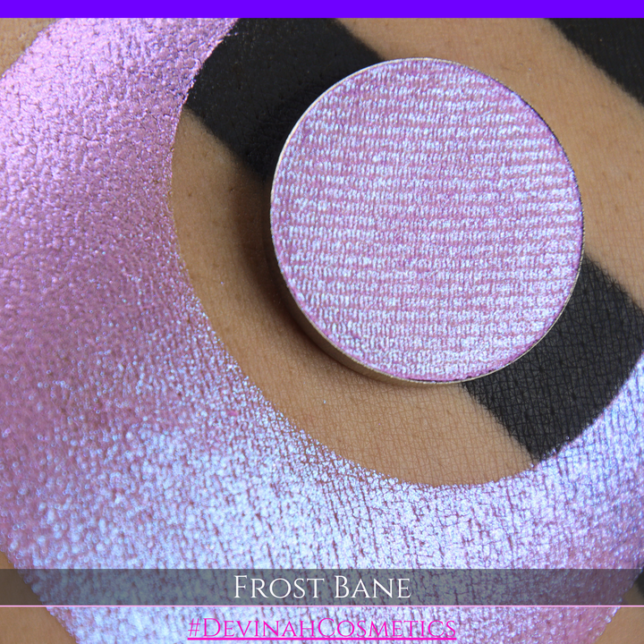 A close-up of skin with a large iridescent pink-blue crescent-shaped makeup swatch and a matching circulareyeshadow pan, with text reading 'FROST BANE' and '#DEVINAH COSMETICS' at the bottom.